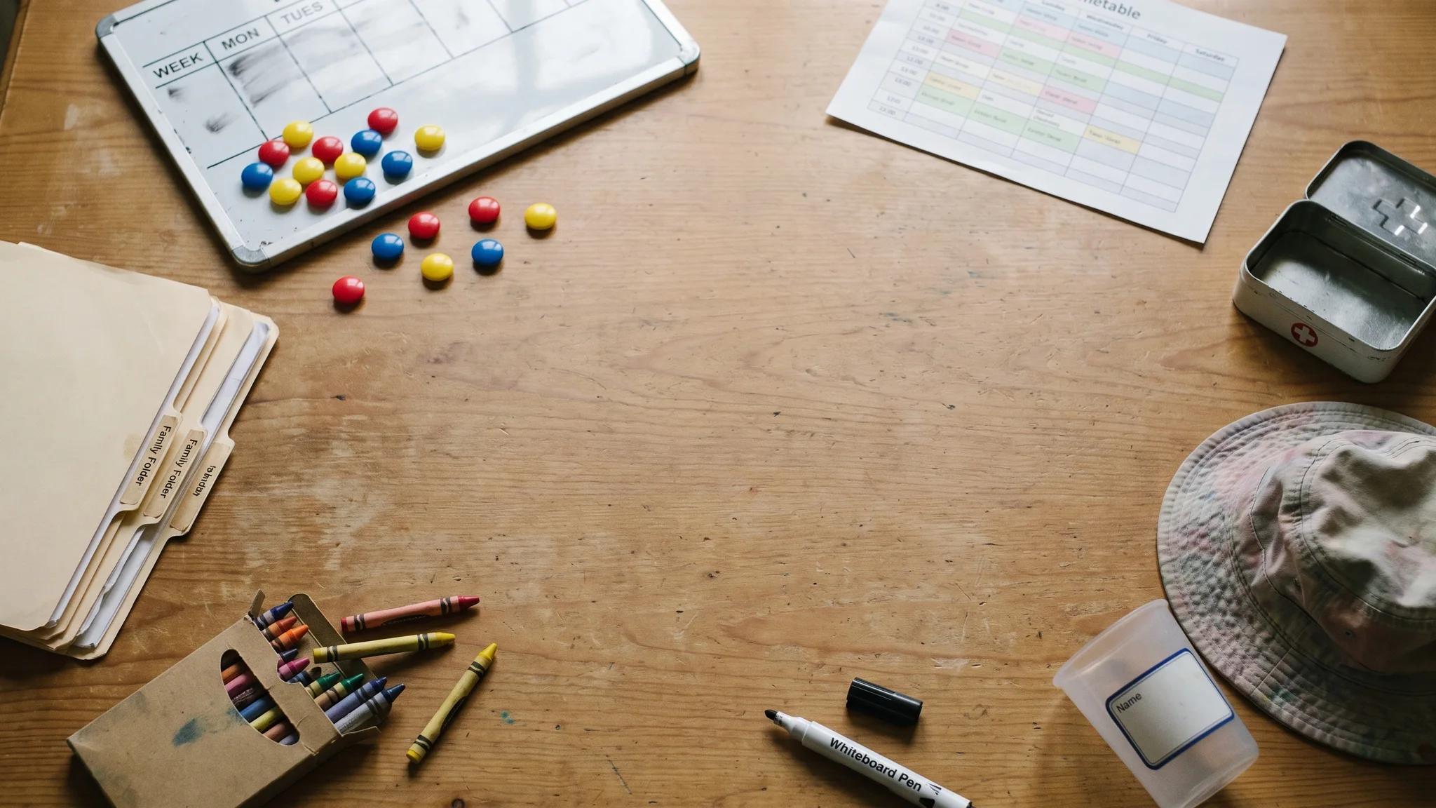 A small whiteboard laid flat on a wooden table showing a half-wiped week grid with red, yellow and blue magnets, beside tabbed family folders, a printed timetable, a box of crayons, a first-aid tin and a named beaker.